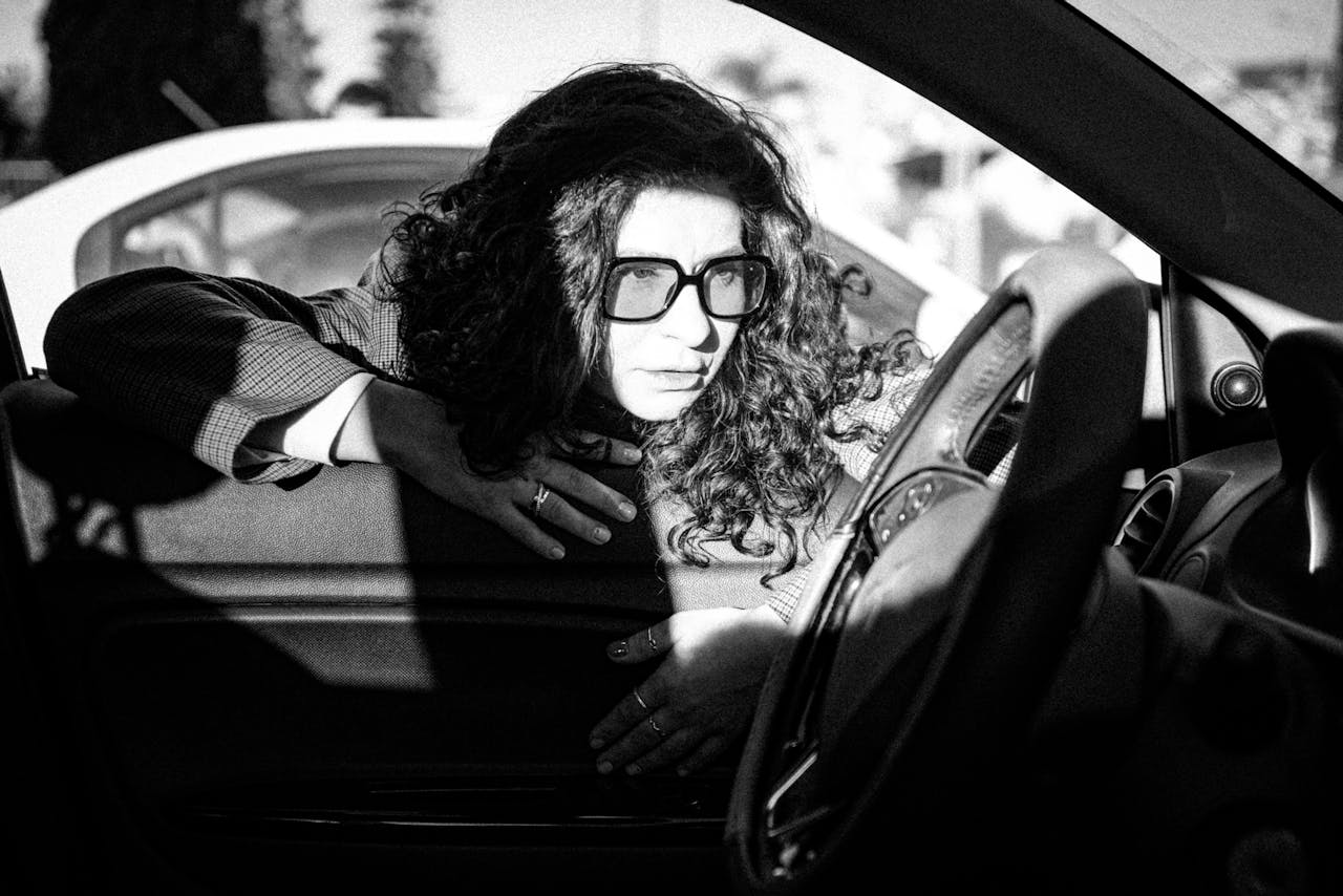 Black and white shot of a woman leaning on a cars interior, wearing sunglasses.